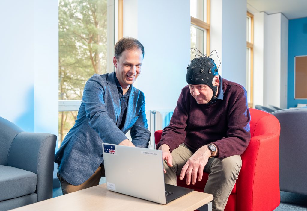 Dr George Stothart is sad next to an older man wearing the EEG headset and both are looking at a laptop on a small table.