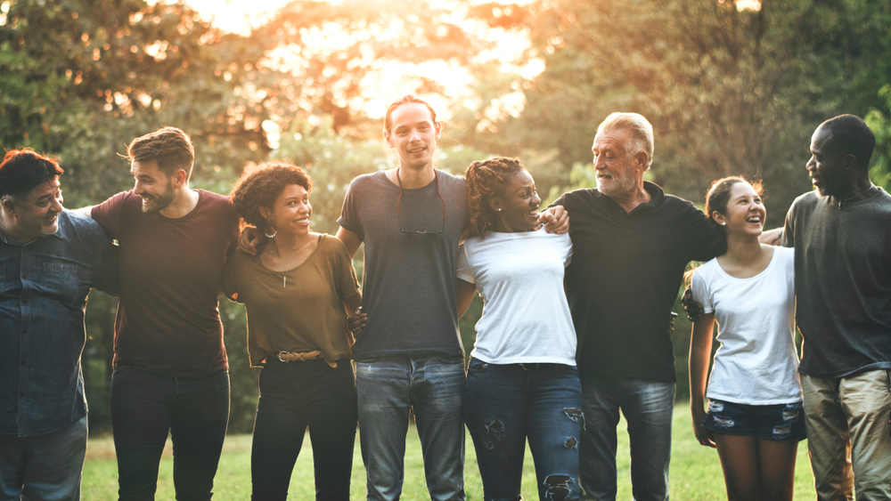 Cheerful,Diverse,People,Huddling,In,The,Park A group of 8 adults of different ages and ethnicity stood next to each other with their arms around each other in a line.