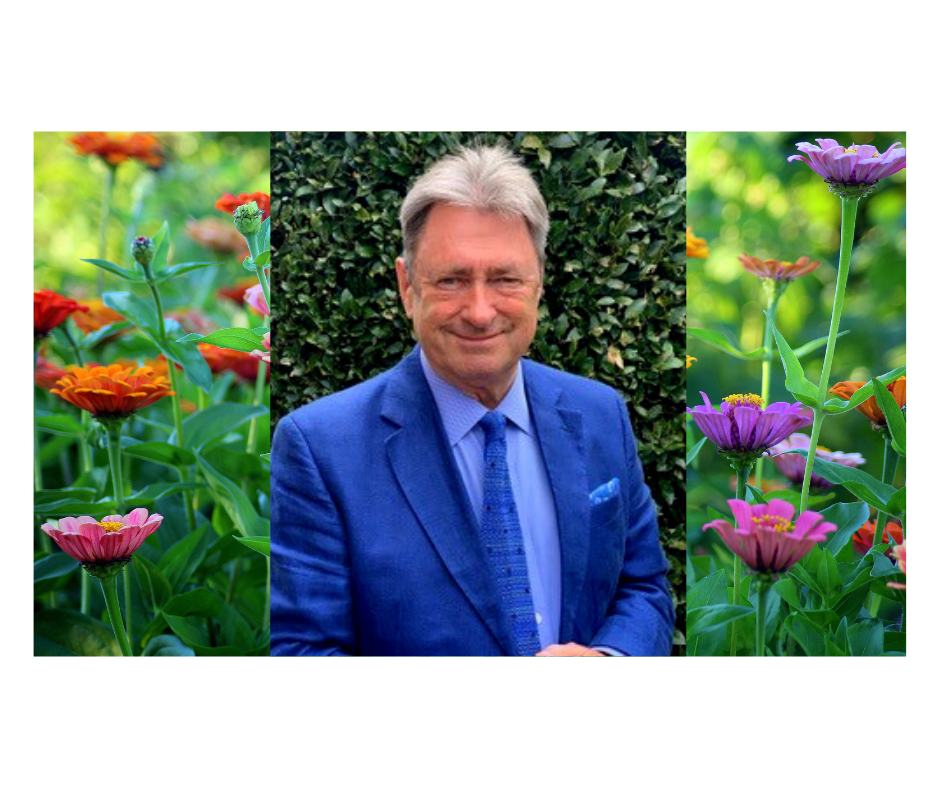 Alan Titchmarsh is in a bright blue suit looking towards you and smiling. Behind him is a photo of brightly coloured wild flowers.