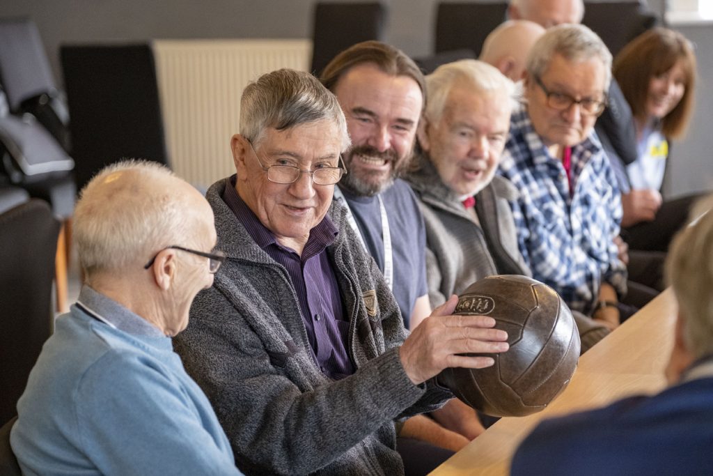 Seven people in their 60s and 70s sat up to a table looking at a man holding a football.