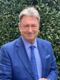 Alan Titchmarch headshot Alan Tichtmarsh is wearing a bright blue suit, shirt and tie. He is stood in front of a bush.