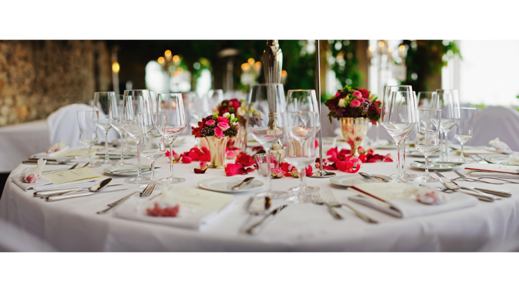 A formal dinning table set with cutlery, different sized wine glasses and napkins. There are also lots of red and pink flowers in vases and red and pink petals scattered in the middle of the table.