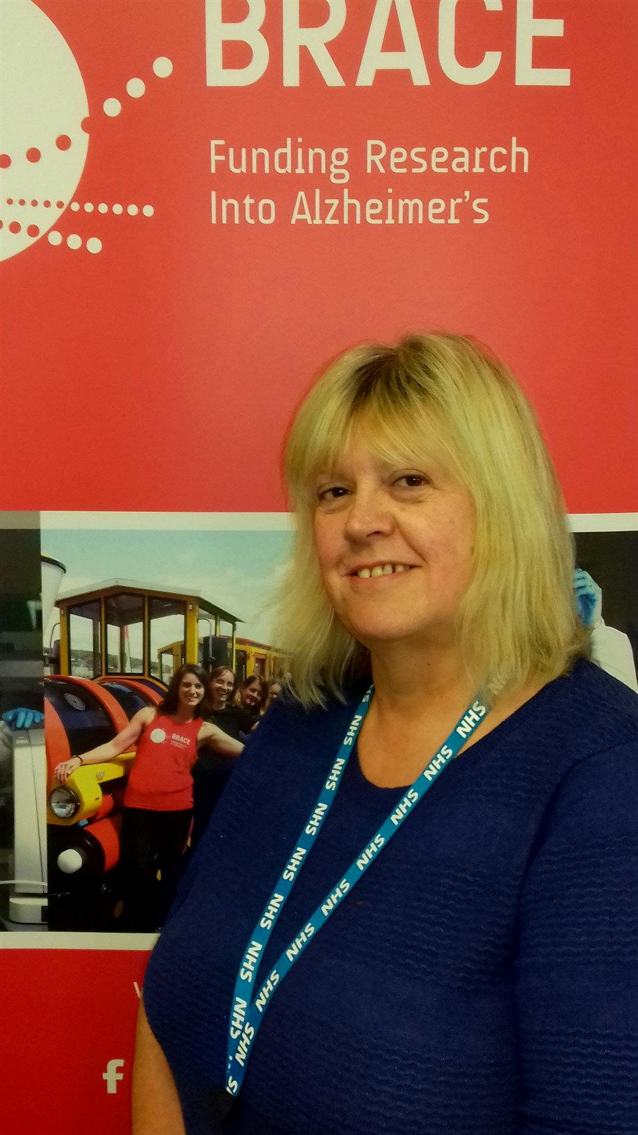 A white woman with blonde hair and a navy top is stood in front of a red BRACE banner