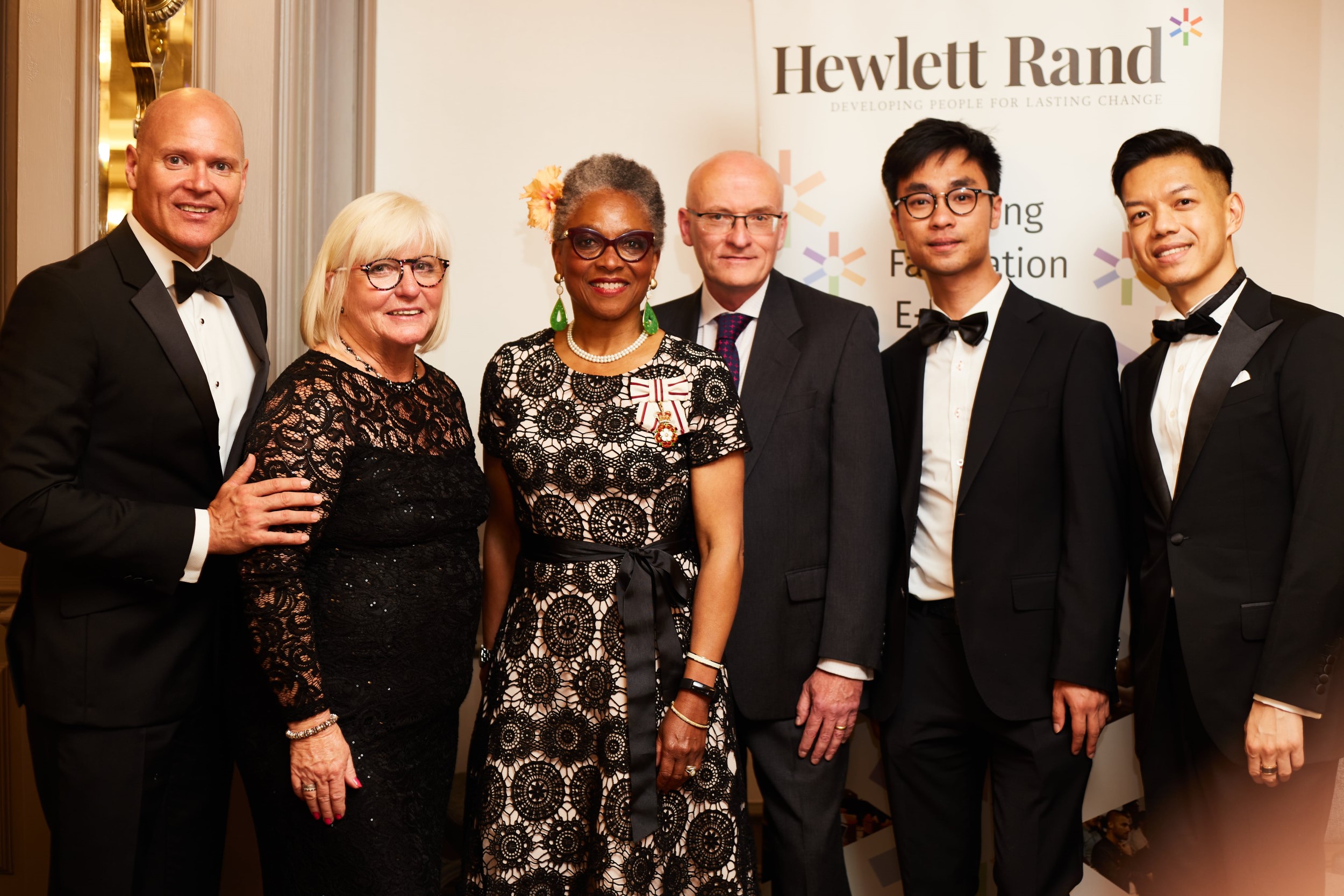 In the middle is Peaches Golding in a black and white dress. To her right are three men in suits. To her left is Richard Lowe in a suit and Jane Dare in a black dress. Behind them is a Hewlett Rand sign.