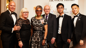 In the middle is Peaches Golding in a black and white dress. To her right are three men in suits. To her left is Richard Lowe in a suit and Jane Dare in a black dress. Behind them is a Hewlett Rand sign.