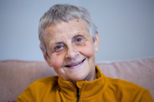 Wendy is a white woman in her 60s. She has short grey hair and green eyes. She is smiling. Wendy is sat on a sofa and is wearing a mustard yellow jumper.