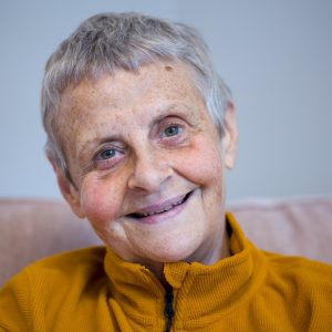 Wendy is a white woman in her 60s. She has short grey hair and green eyes. She is smiling. Wendy is sat on a sofa and is wearing a mustard yellow jumper.