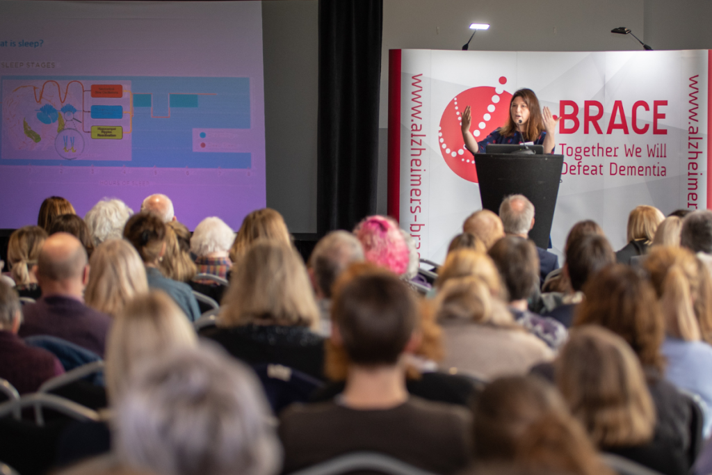 A conference room of people are sat down facing a stage, where a speaker is giving a talk. The speaker is a woman in her late 30s with dark hair, she is motioning with her hands upwards. Behind her is a large BRACE banner.