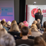 A conference room of people are sat down facing a stage, where a speaker is giving a talk. The speaker is a woman in her late 30s with dark hair, she is motioning with her hands upwards. Behind her is a large BRACE banner.