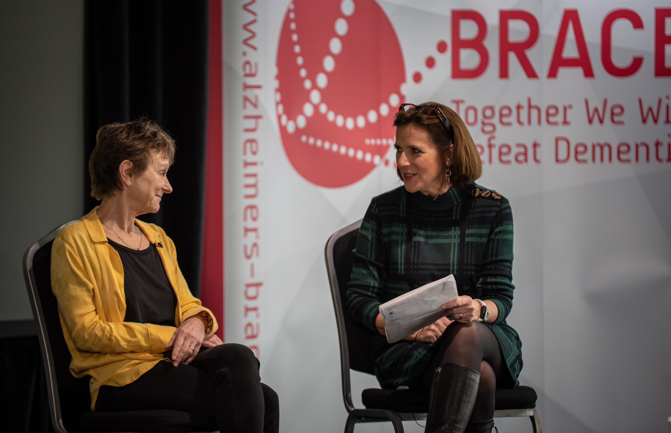 A middled aged woman is sat on a stage. She is wearing a bright yellow cardiagan. To her right is another middle aged woman in a dark green dress, she is also sat on a chair. They are facing each other and both smiling in agreement.