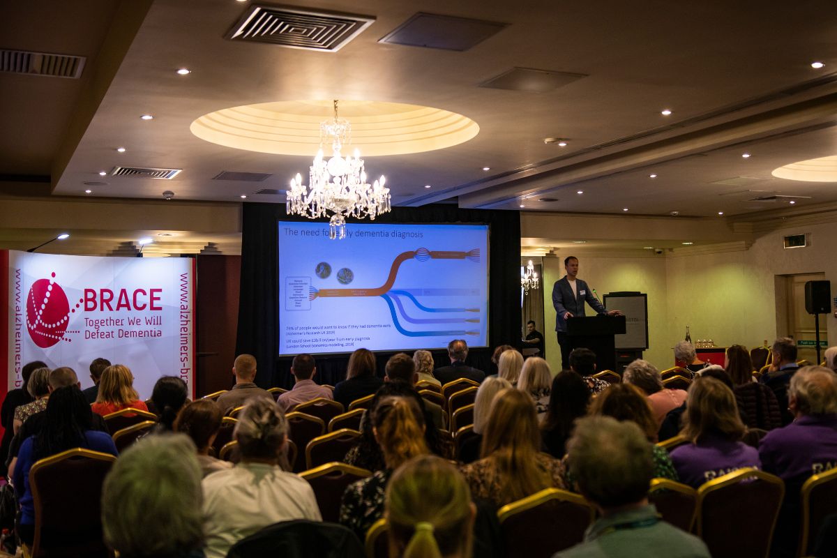 A photo of a large conference room. The audience are sat facing a stage and watching speaker, Dr George Stothart, give his talk. He has a big screen next to him with slides on.