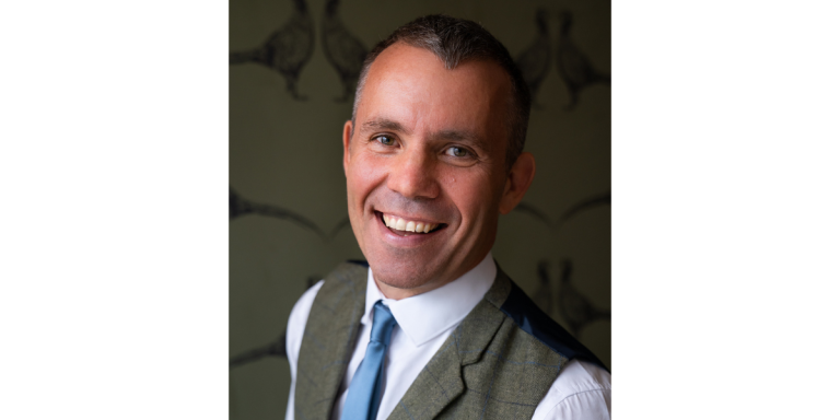 Richard Higgs is a white middle aged man, wearing a shirt, blue tie and a grey waistcoat. He is smiling and looking forward.