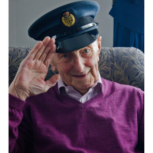 An older man sat with an army hat on saluting.