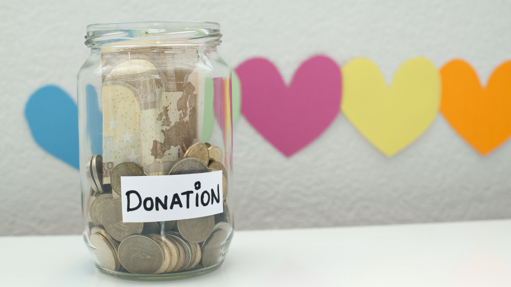 A glass jar filled with coins, with the word donations on is on a table. Behind are paper hearts.