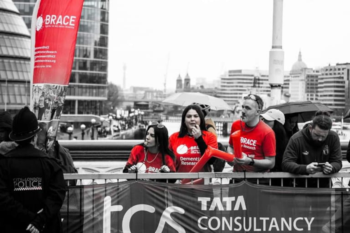 Three people in BRACE red t-shirts are stood on Tower Bridge with a BRACE banner during the London Marathon 2023.