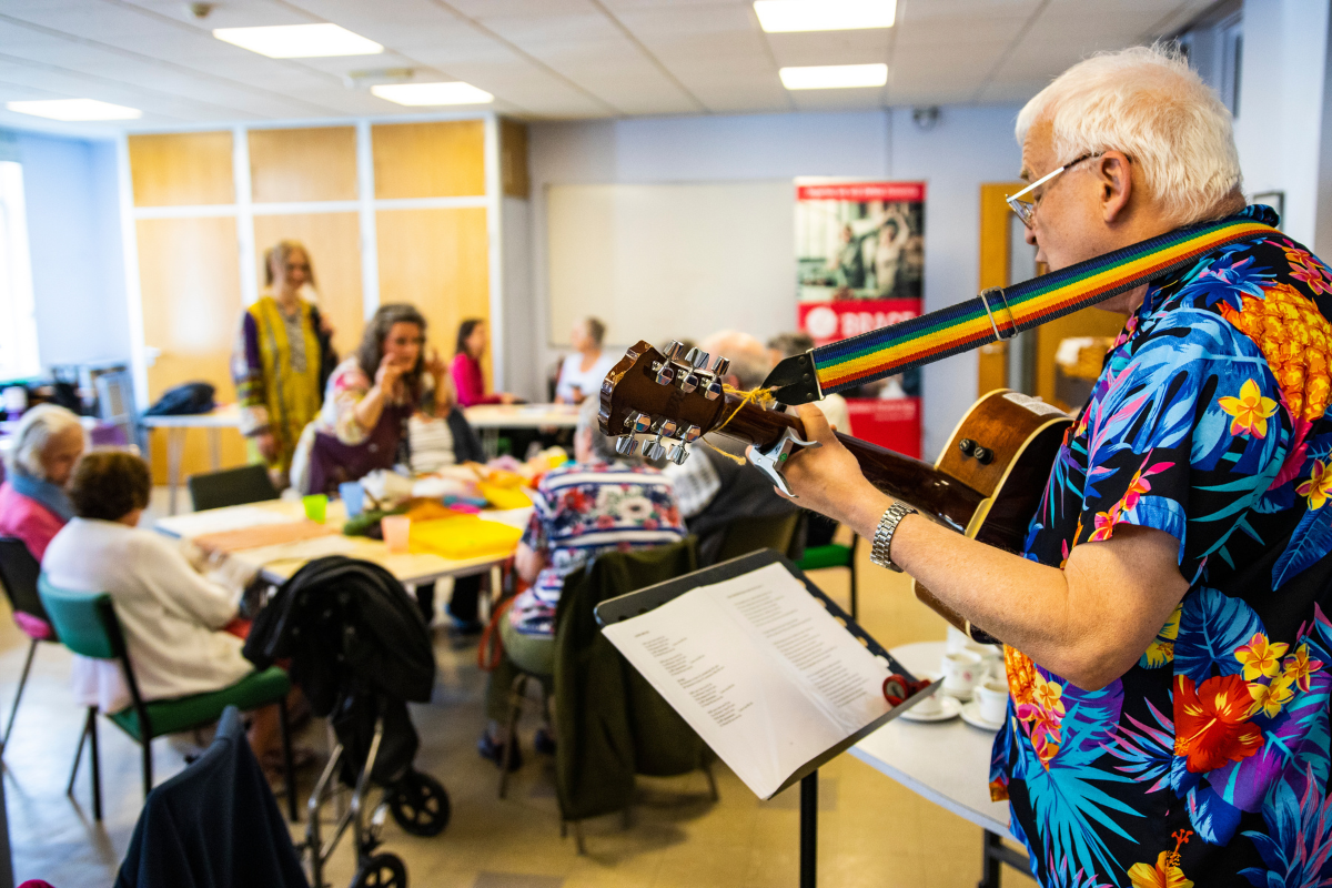 A man with white hair and an Hawaiian shirt is playing a guitar to lots of people sat down to tables.