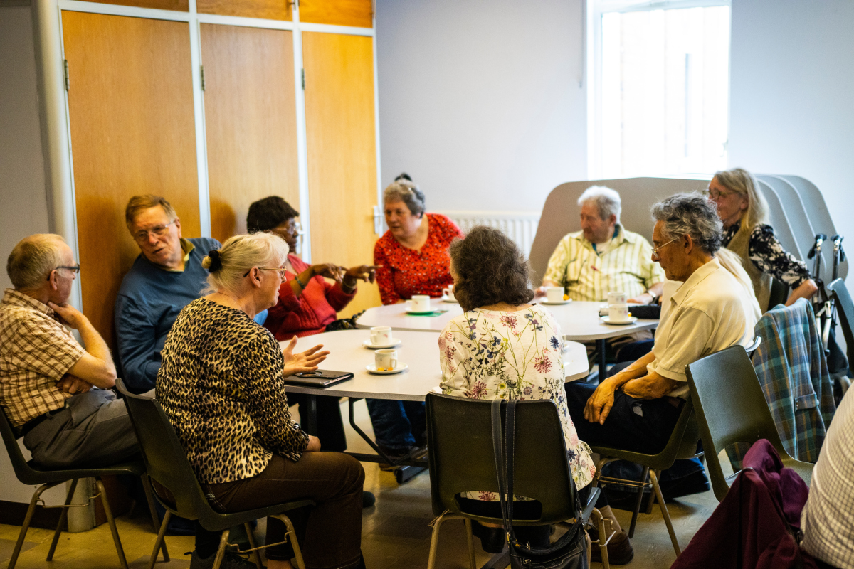 A group of people are sat around a table having conversations and drinking tea / coffee.