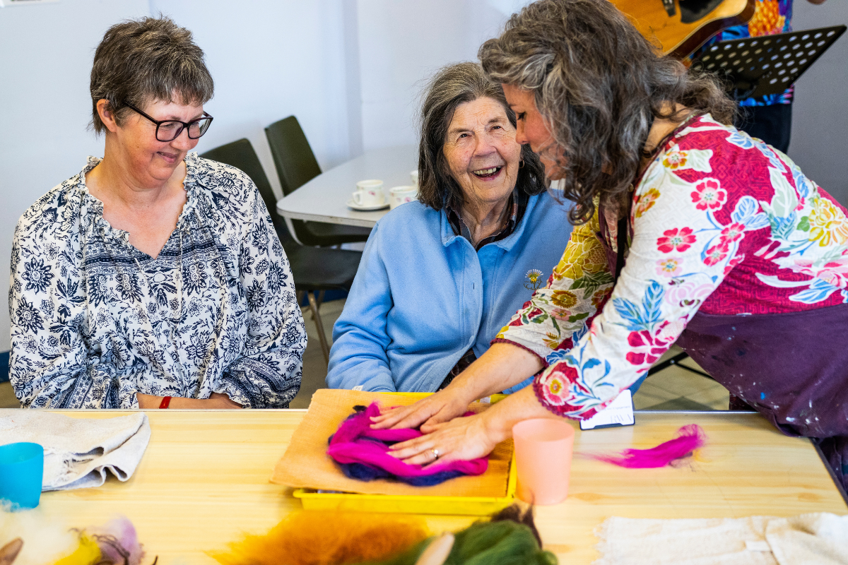 A woman sat at a table smiles as she is taught how to make felt art.
