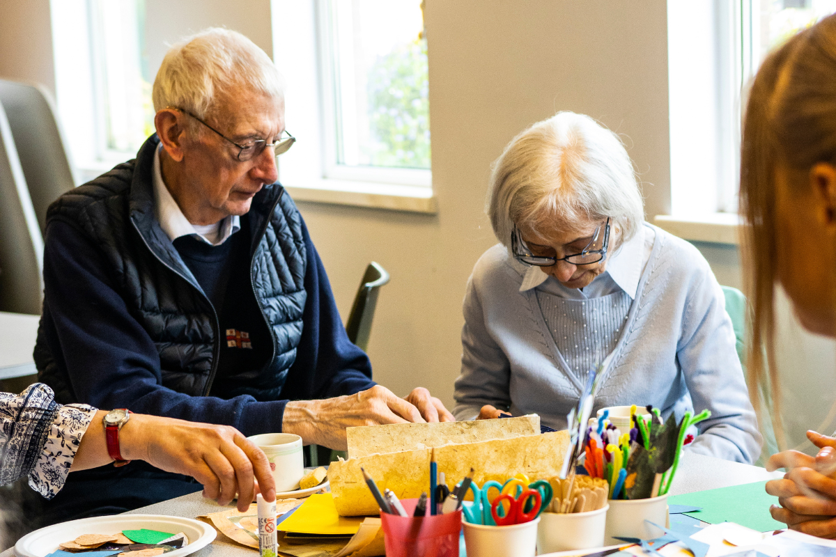 A man and a women sat at a table making art with mugs of tea. The table is covered in equipment; glue, scissors, pencils and paper.