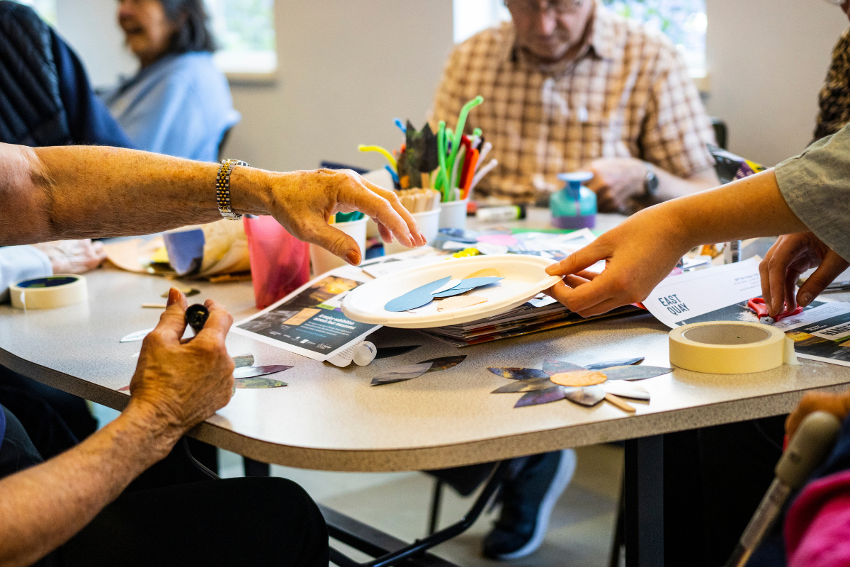 A woman holds a plate to someone sat at a table so they can take materials to make art.