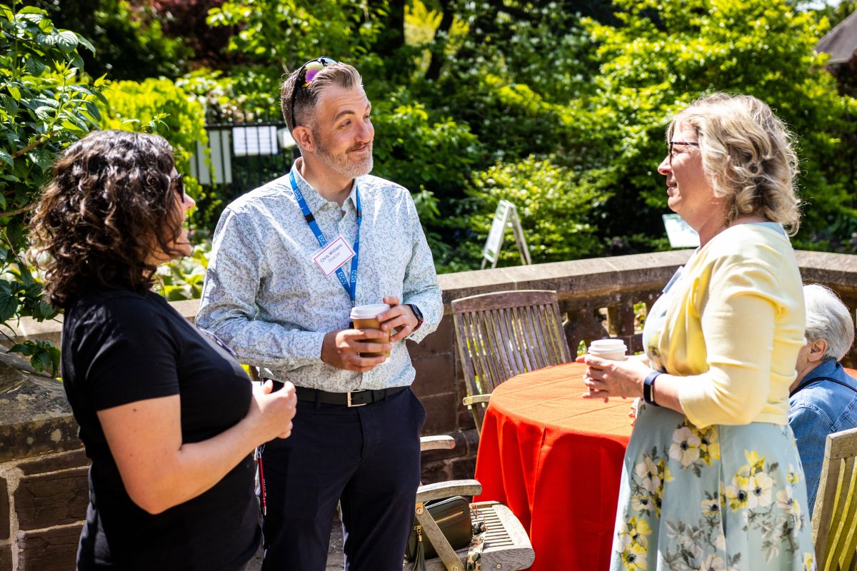 A man and two women have a conversation while holding cups of tea in the sunshine.