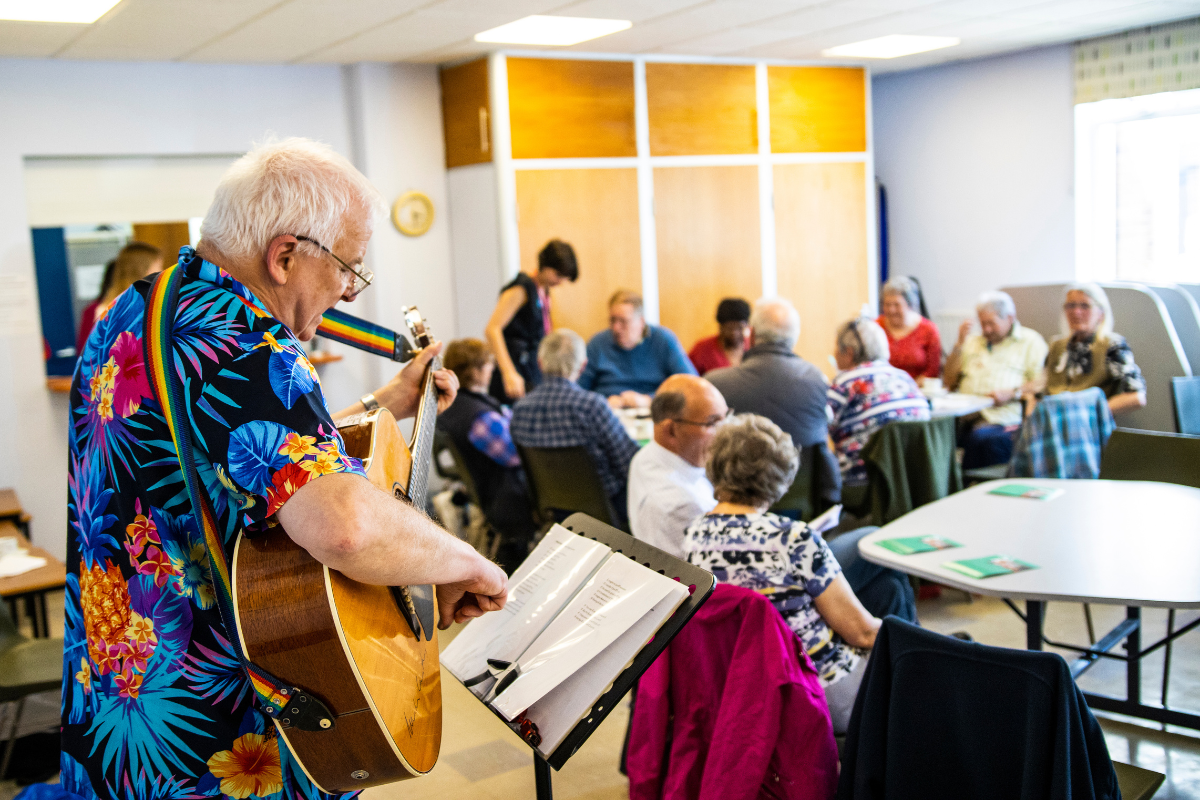 A man with white hair and an Hawaiian shirt is playing a guitar to lots of people sat down to tables.