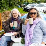 Two women are sat on a bench drinking tea and holding plates with cake on.