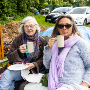Two women are sat on a bench drinking tea and holding plates with cake on.