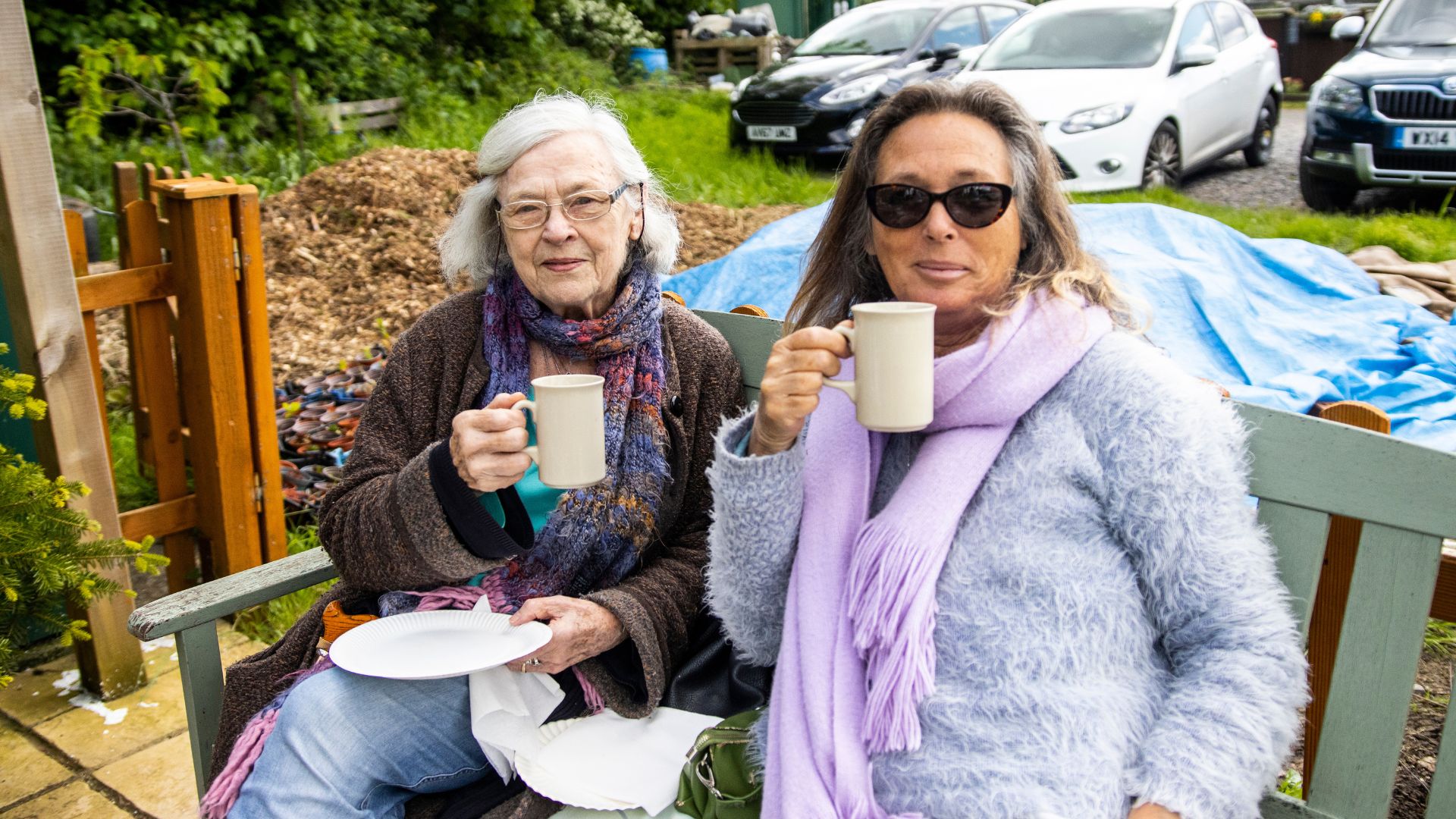 Two women are sat on a bench drinking tea and holding plates with cake on.