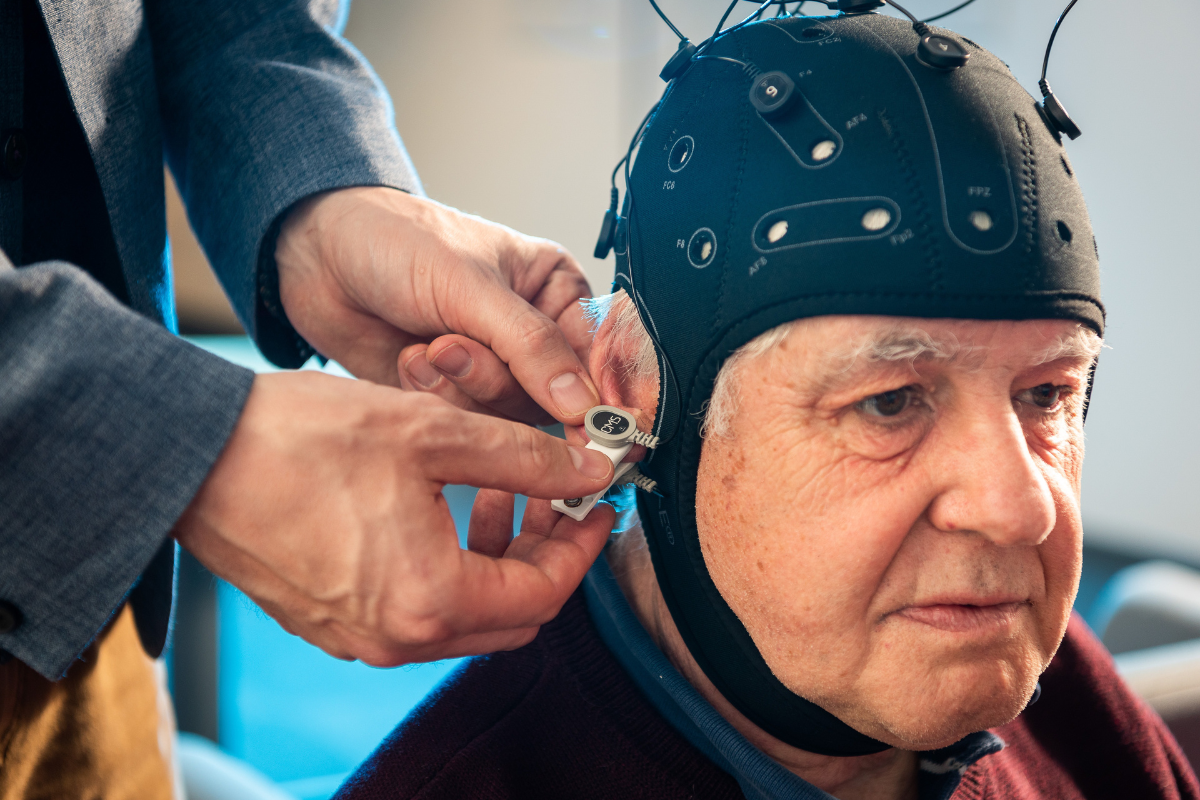An older man has a black cap covering his head. The cap is the Fastball test. It has some wires coming out of the top. A man's hands can be seen attaching a clasp to one of the wires.