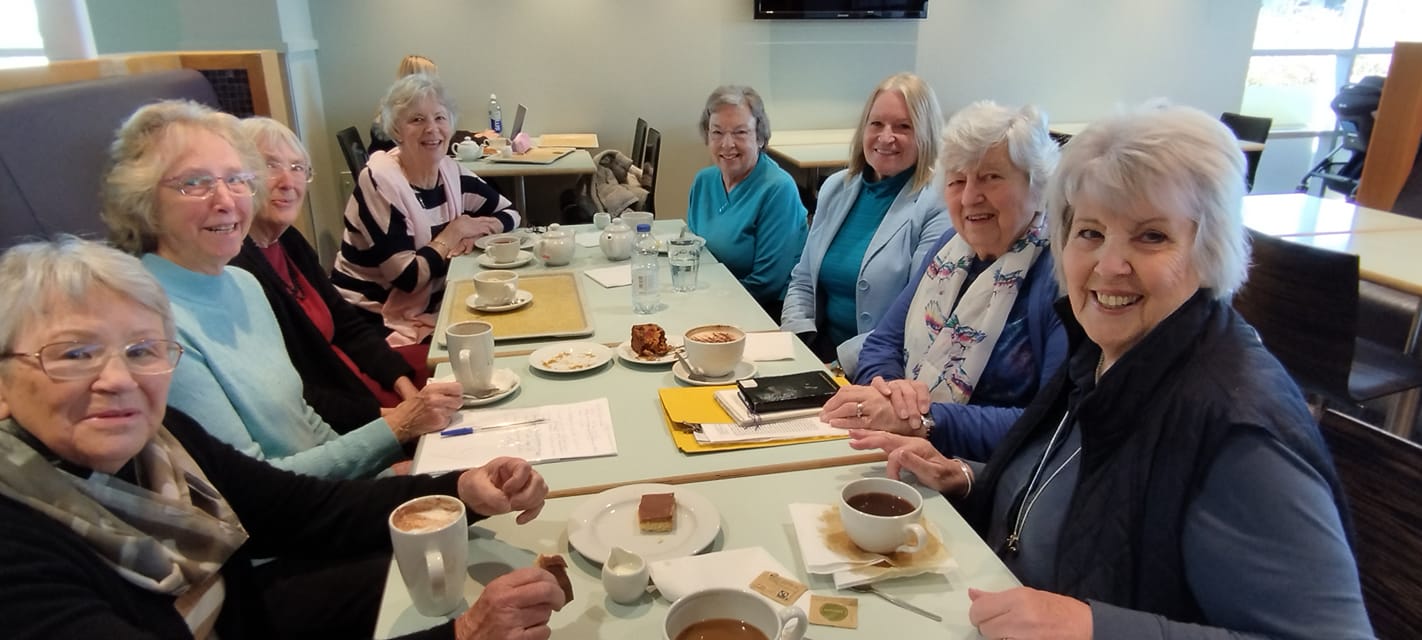 A group of women sit around a table with mugs of hot drinks and cakes.