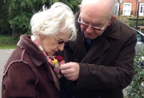 An older couple together. The husband is adjusting a necklace which is round his wife's neck.