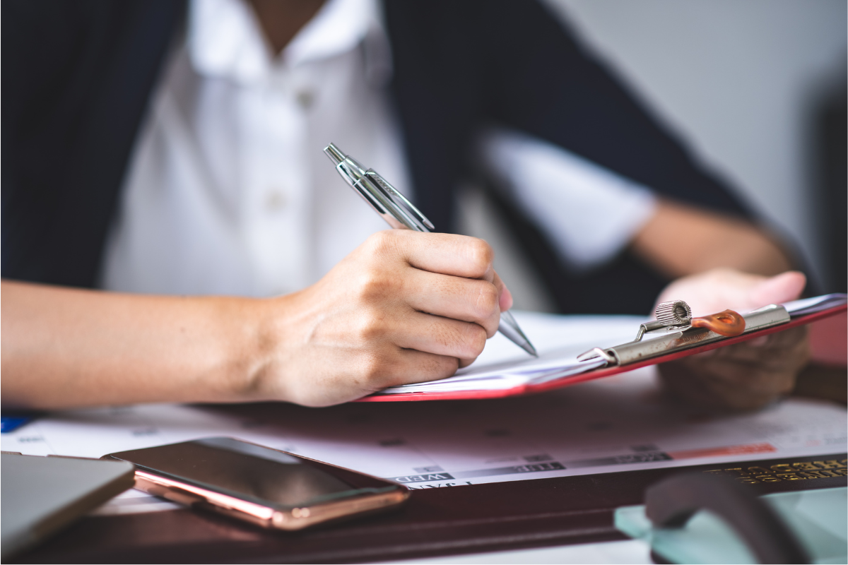 A persons hand who is writing on paper and a red clipboard with a pen.