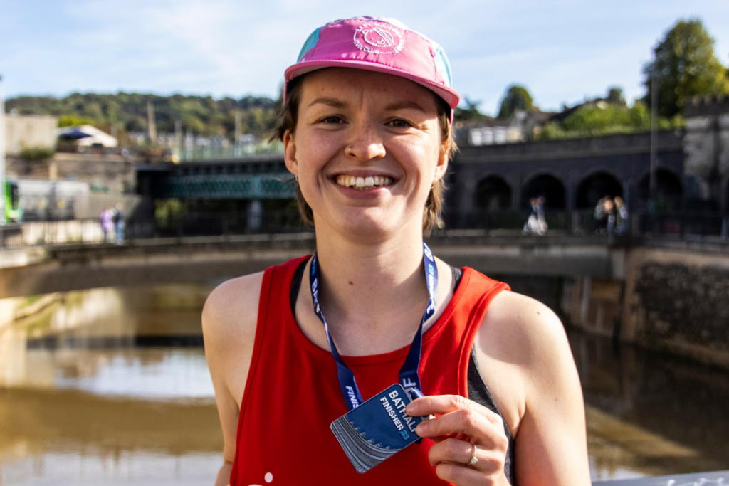 A woman wearing a red BRACE vest is smiling holding up a Bath Half Finisher 23 medal.