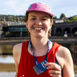 A woman wearing a red BRACE vest is smiling holding up a Bath Half Finisher 23 medal.