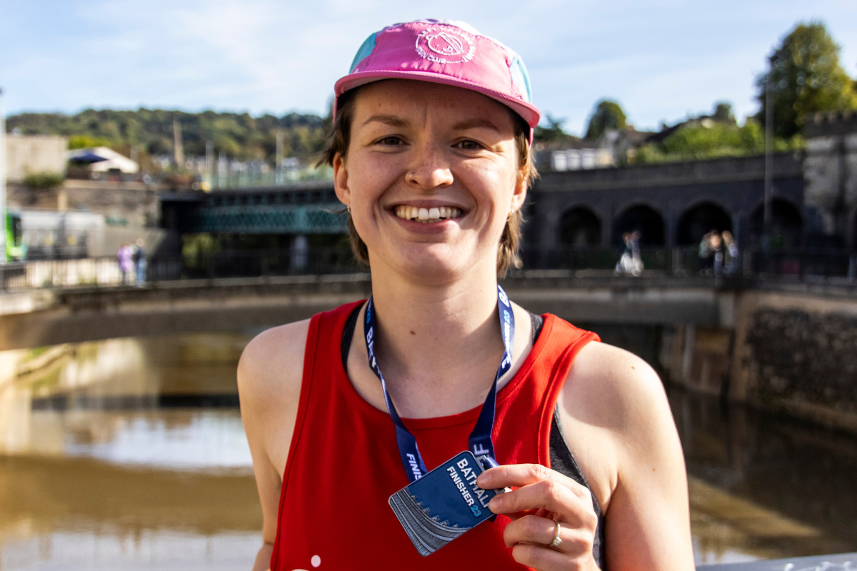 A woman wearing a red BRACE vest is smiling holding up a Bath Half Finisher 23 medal.