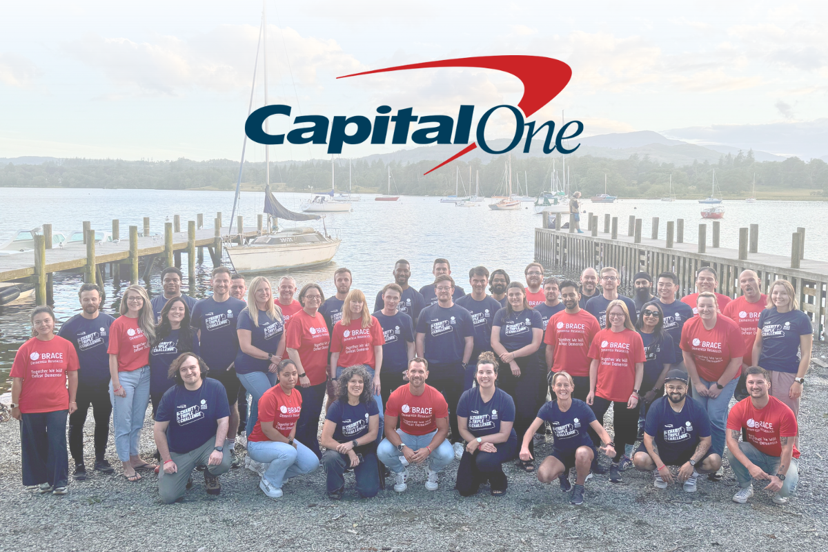 Capital One (1) A large group of people wearing BRACE charity t-shirts in red and navy t-shirts pose by a lakeside with wooden docks and sailboats in the background. The 'Capital One' logo is displayed above them.