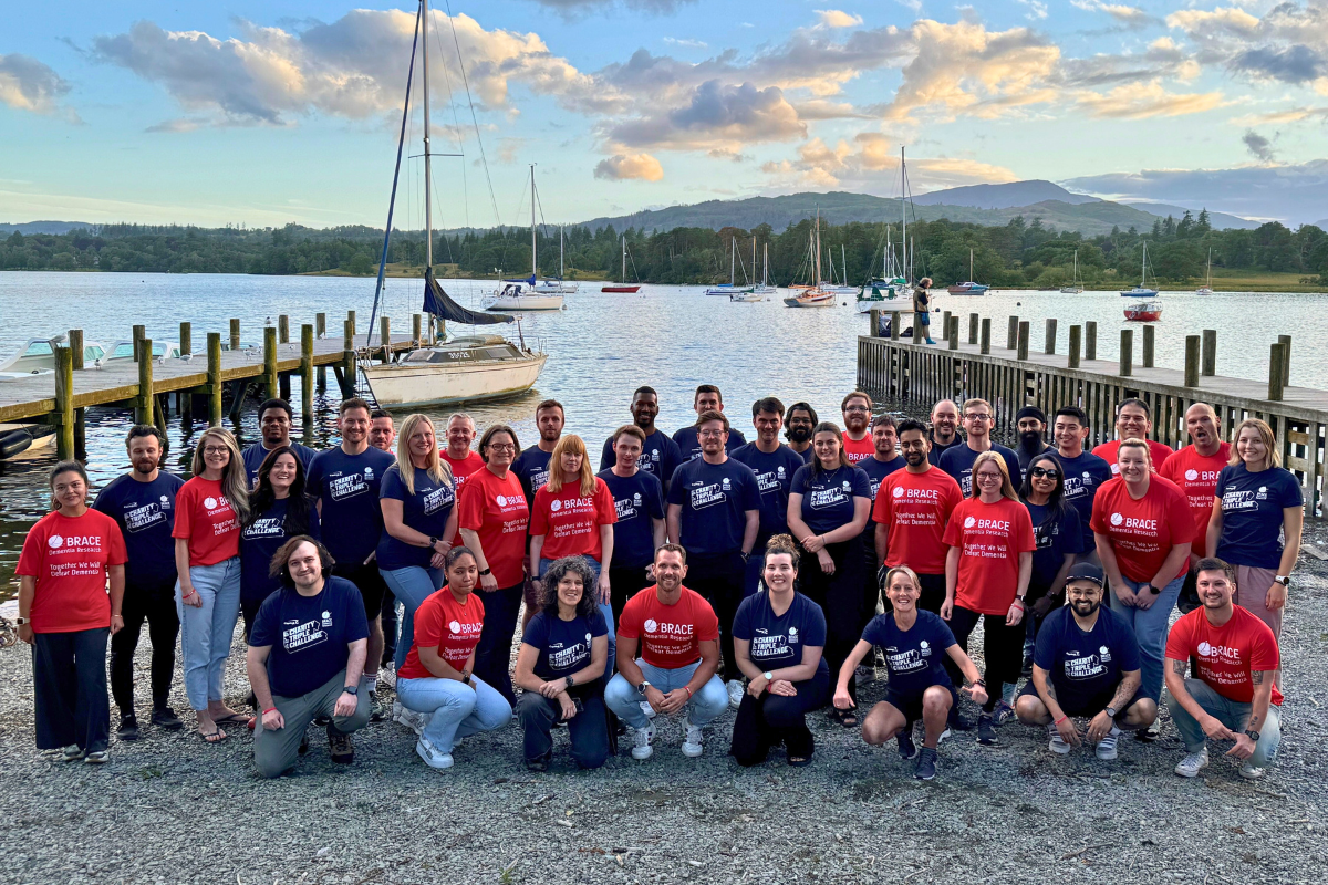 A large group of people wearing BRACE charity t-shirts in red and navy t-shirts pose by a lakeside with wooden docks and sailboats in the background.