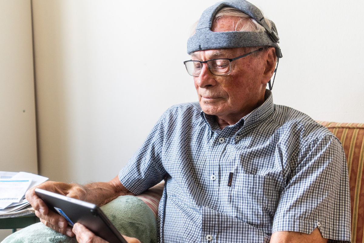 An older man is sat on a sofa with the Fastball headset on and looking at the tablet.