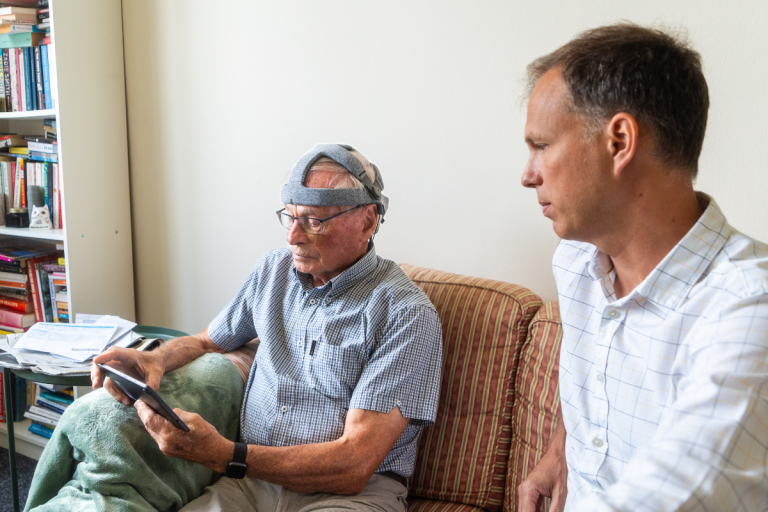 Dr Stothart and a research volunteer are sat on a sofa. The volunteer has the Fastball cap on and is taking the test.