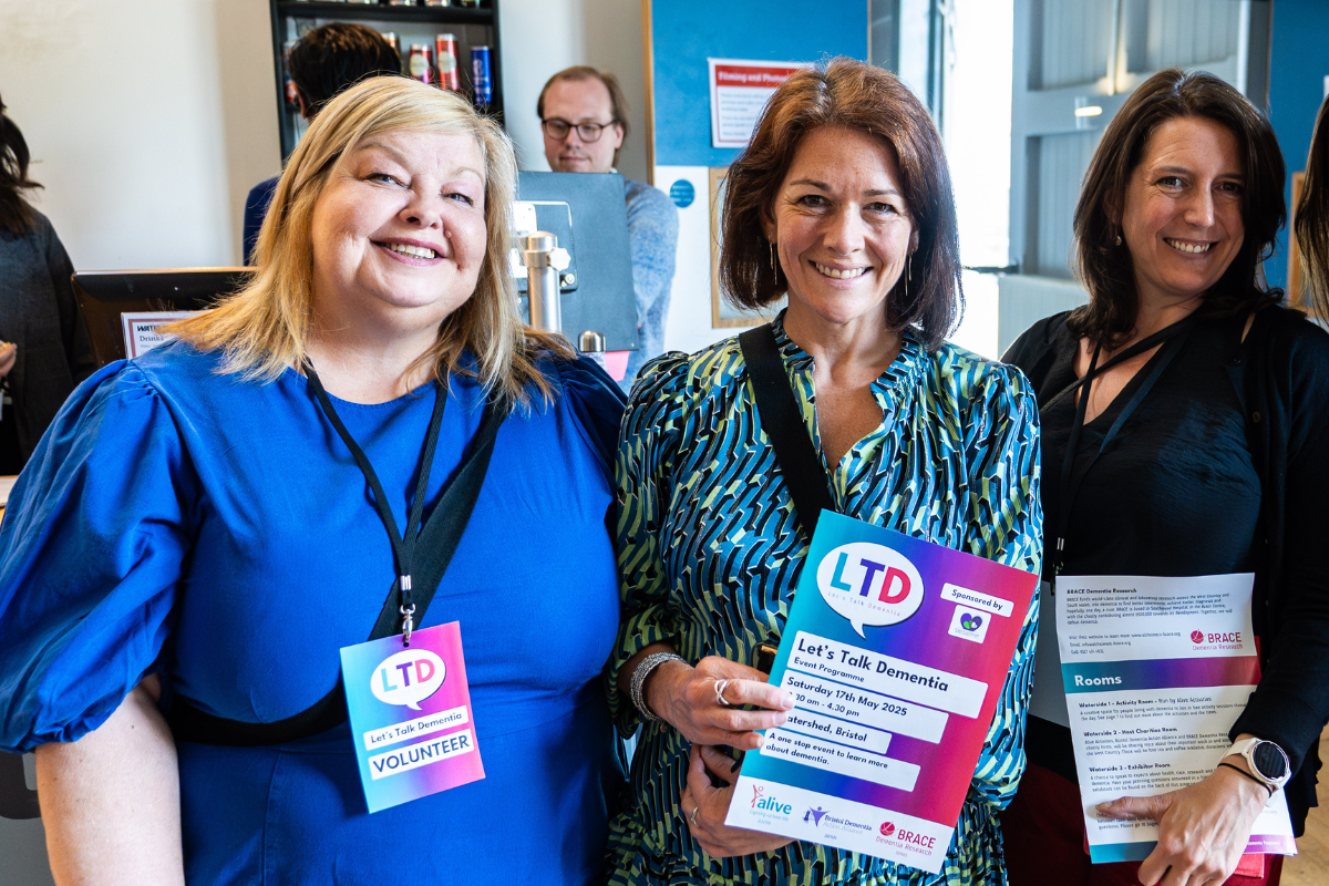 Three women are smiling, holding Let's Talk Dementia programmes and have volunteer tags round their necks.
