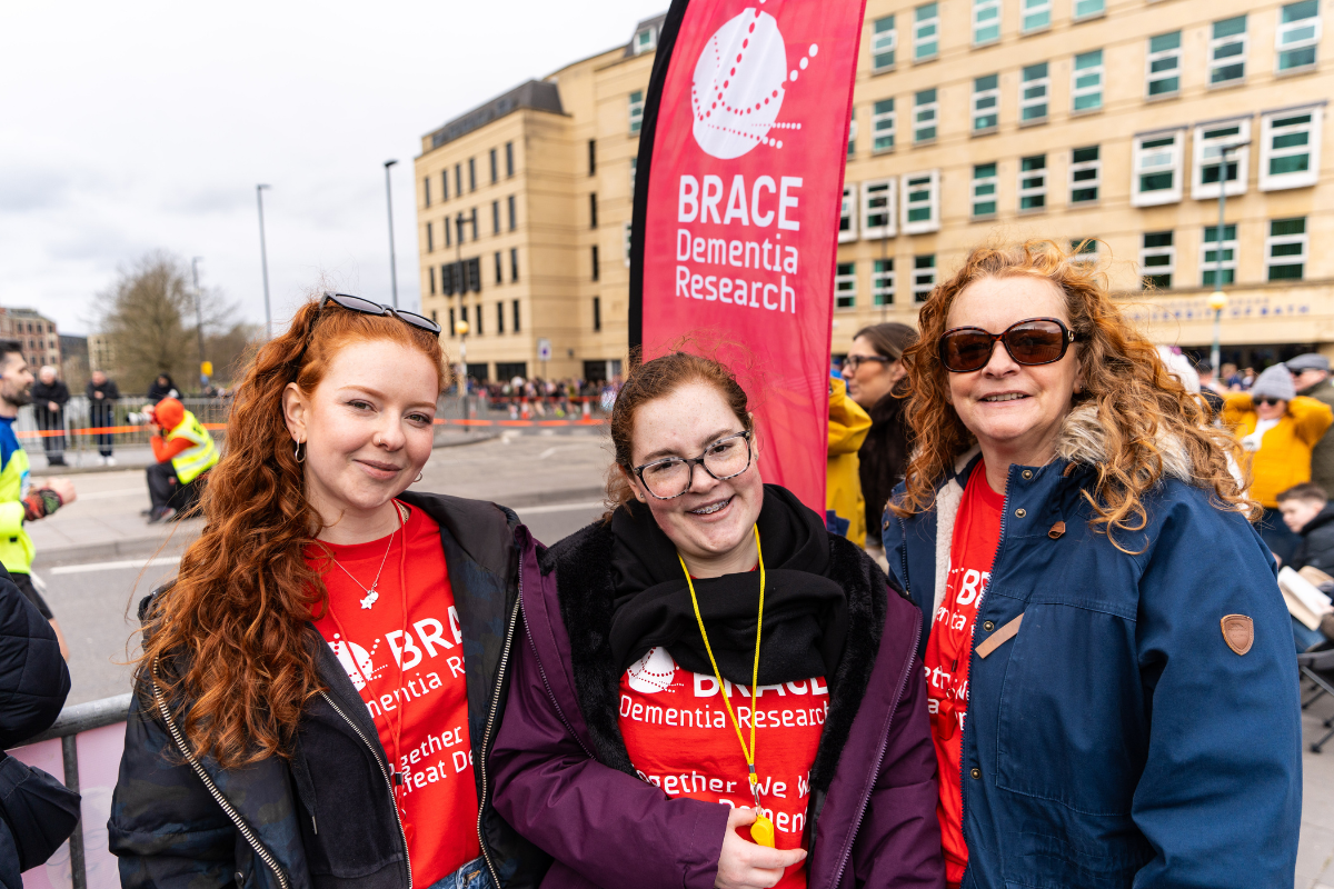 Three people wearing red BRACE t-shirts, standing in front of a red BRACE flag at the Bath Half Marathon cheer point.