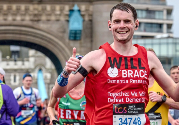 A group of people are running across Tower Bridge at the London Marathon. A man is wearing a red BRACE vest.