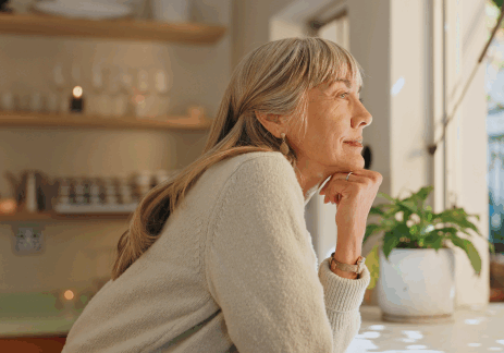 A white woman with grey hair, who is about 60 years old, is looking out of a window.