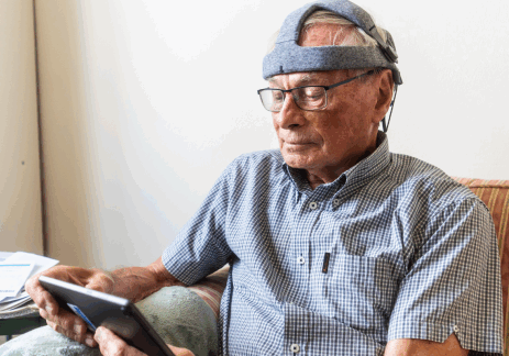 An older man is sat on a sofa with the Fastball headset on and looking at the tablet.