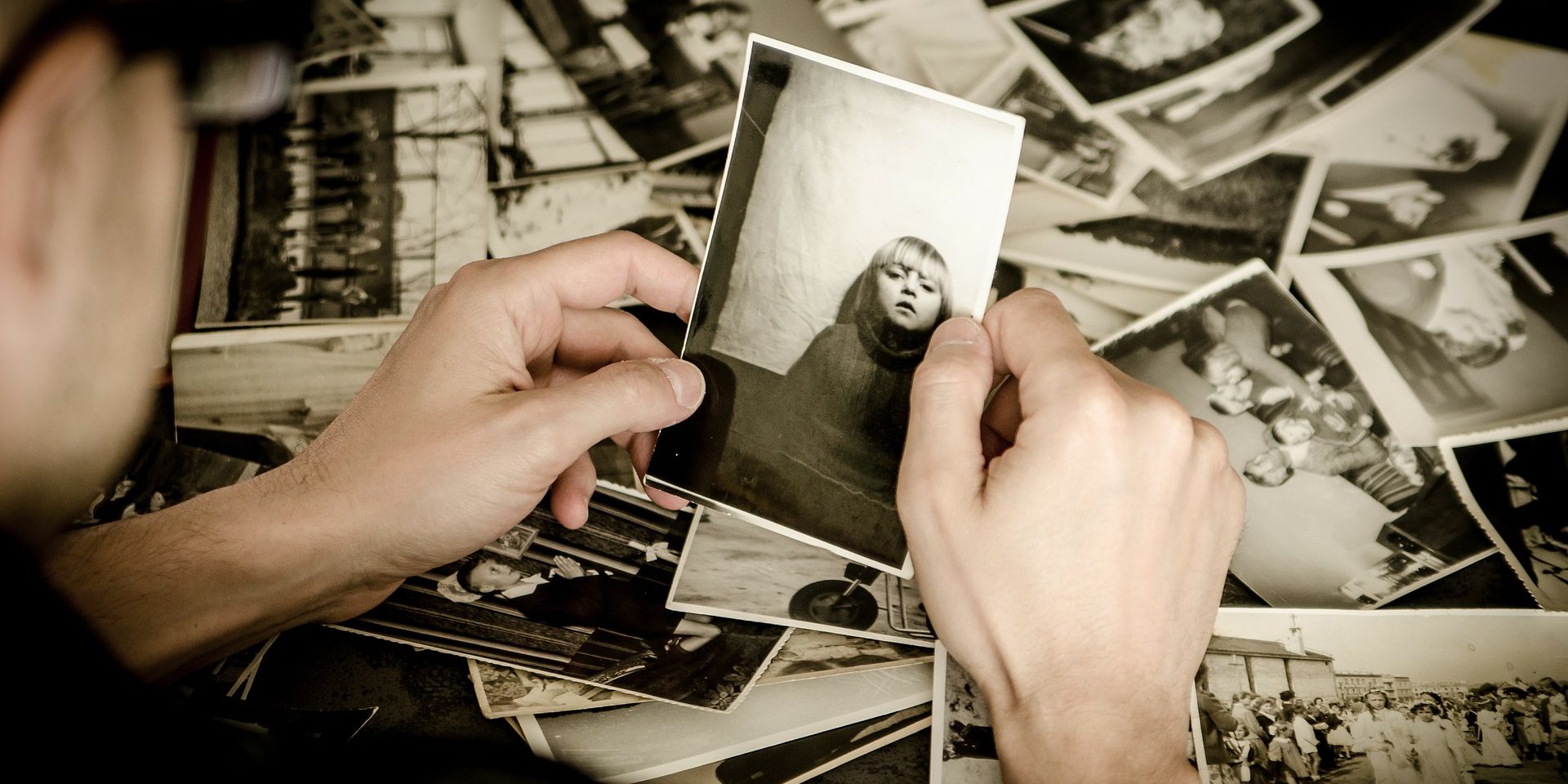Black and white photos laid on table. The back of a person's head, with their hands holding a photo.