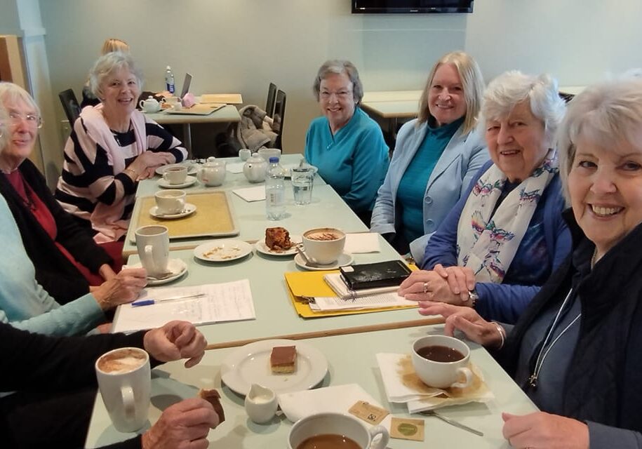 A group of women sit around a table with mugs of hot drinks and cakes.
