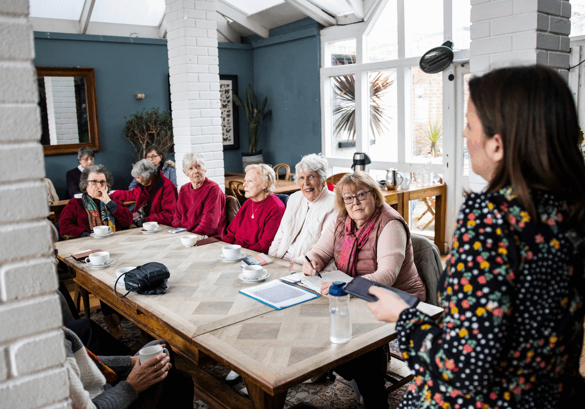 A woman is standing and speaking to a group of people who are sat around a table.