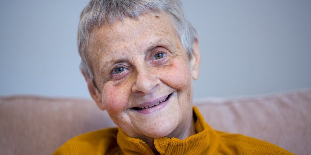 Wendy Mitchell Wendy is a white woman in her 60s. She has short grey hair and green eyes. She is smiling. Wendy is sat on a sofa and is wearing a mustard yellow jumper.