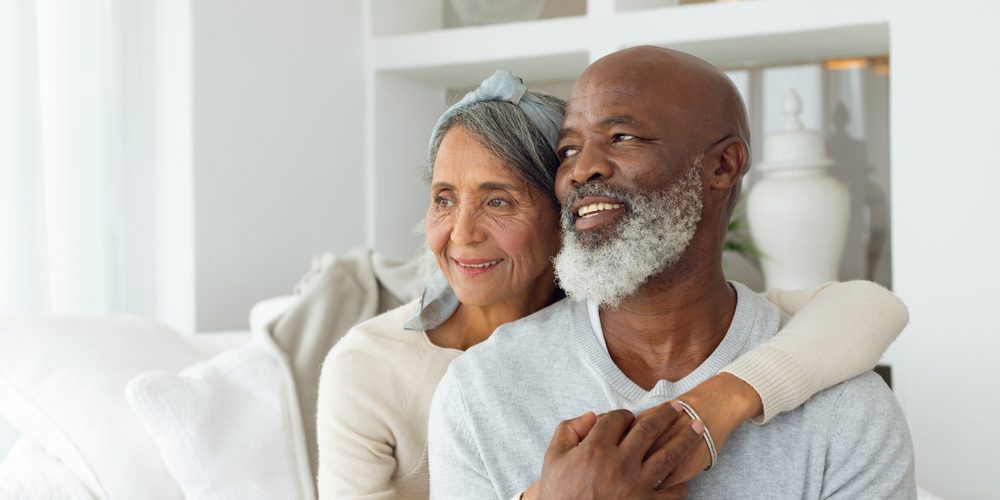Senior couple sitting on a sofa Woman with light brown skin hugging her partner with dark brown skin, he has a white beard. They are sat on a sofa.